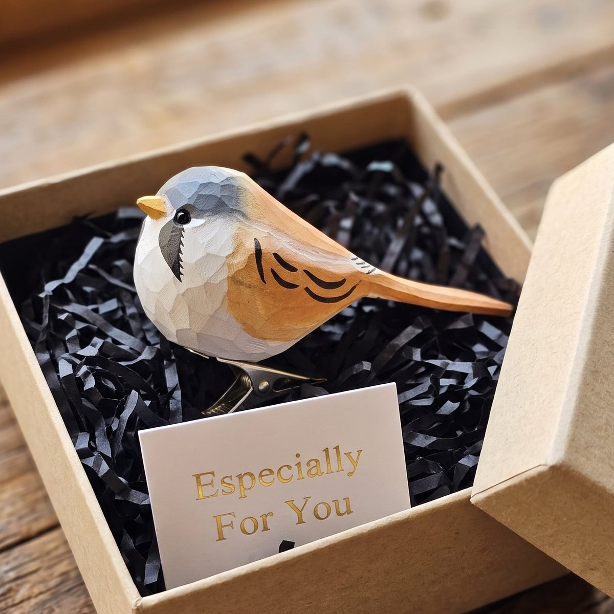 A hand-carved wooden Bearded Tit clip-on bird ornament resting inside an open kraft paper gift box filled with black shredded paper, featuring a slate-grey head, a distinctive black moustache mark below its dark eye, a bright yellow beak, and tawny-orange wings with black-and-white accents, alongside a white card reading "Especially For You" placed in the foreground on a rustic wooden surface.