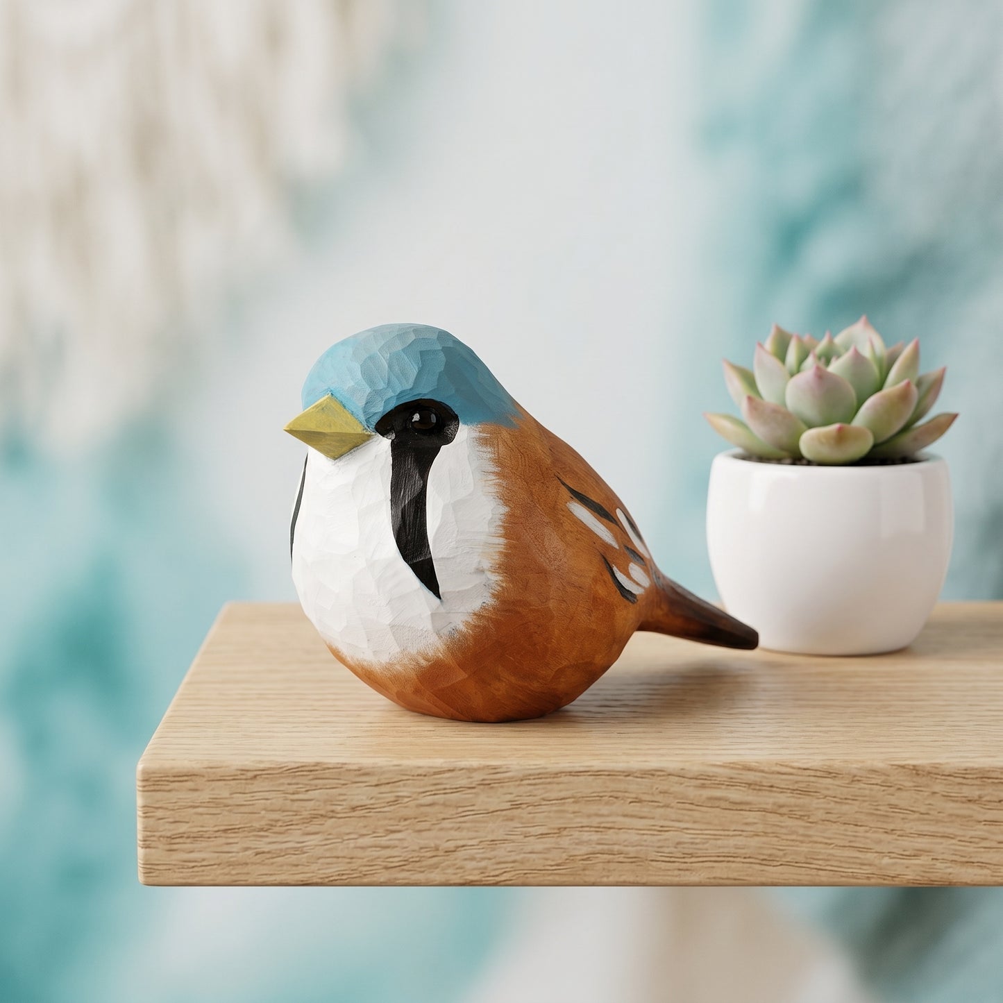 A decorative Bearded reedling figurine with a geometric, low-poly style, featuring a blue head, white chest, brown body, and yellow beak, sitting on a light wood shelf next to a small potted succulent.