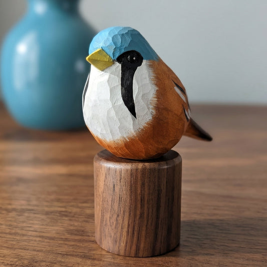 A hand-carved wooden Bearded reedling figurine with a blue, white, and brown painted finish, mounted on a cylindrical wooden base and displayed on a wooden tabletop with a blurred blue vase in the background.
