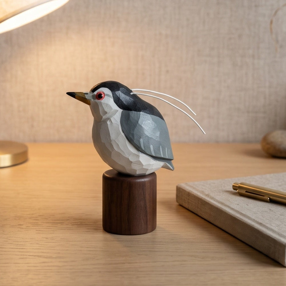 A small geometric Black-crowned Night Heron figurine with a dark gray and white body, red eye, and long white tail feathers, displayed on a dark wooden base on a wooden desk next to a lit brass lamp and a notebook.