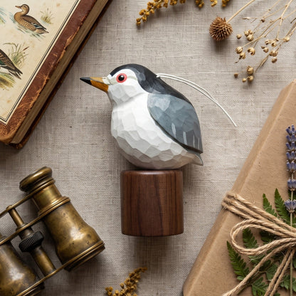 A geometric Black-crowned Night Heron decorative object with a dark gray and white body, yellow beak, and red eye, mounted on a round wooden base. It is styled in a flat lay with dried flowers, an old bird book, brass binoculars, and a wrapped gift on a linen surface.