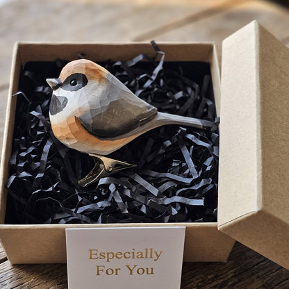 A hand-carved wooden Black-Throated Bushtit clip-on bird ornament resting inside an open kraft paper gift box filled with black shredded paper, featuring a warm chestnut cap, a distinct black throat patch, pale grey wings, and a long tail, alongside a white card reading "Especially For You" placed in the foreground on a rustic wooden surface.