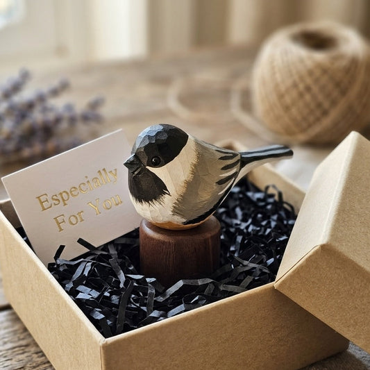 A small painted Black-capped chickadee figurine perched on a wooden stand, displayed inside a kraft gift box with black shredded paper and a card that reads 'Especially For You.' The box is open and placed on a rustic wooden table, with a spool of twine and soft-focus decor in the background.