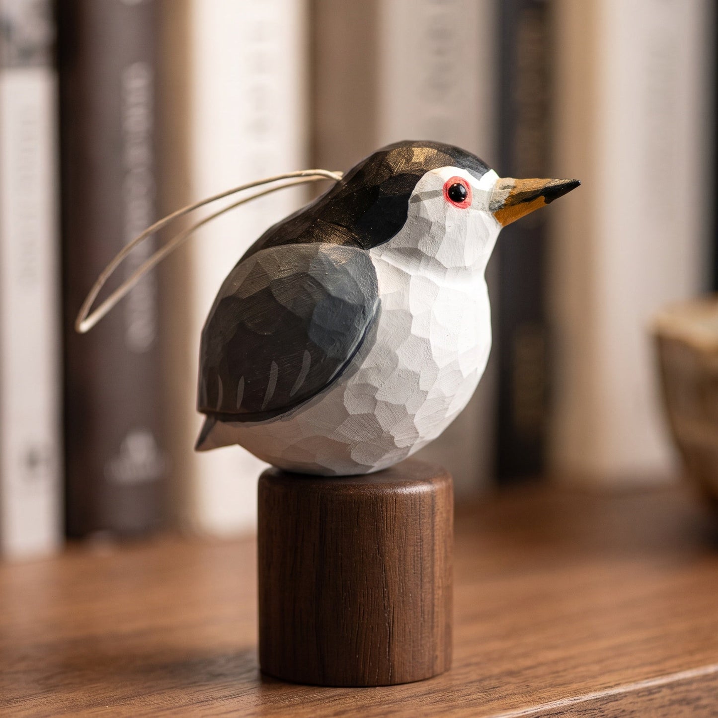 A small carved Black-crowned Night Heron figurine with a white and dark gray body, yellow beak, and red eye, mounted on a cylindrical wooden base on a wooden surface with blurred books in the background.