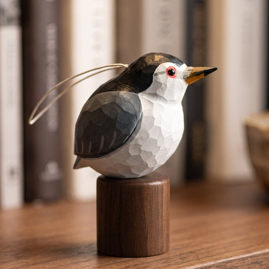 A small carved Black-crowned Night Heron figurine with a white and dark gray body, yellow beak, and red eye, mounted on a cylindrical wooden base on a wooden surface with blurred books in the background.