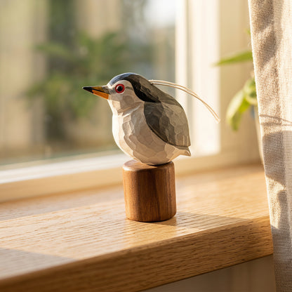 A geometric, faceted Black-crowned Night Heron figurine in gray, white, and black with a red eye and yellow beak, mounted on a small wooden cylinder and displayed on a windowsill in warm natural light.