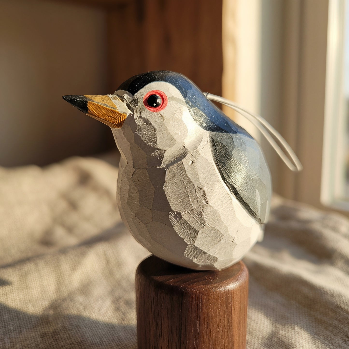 A handcrafted wooden Black-crowned Night Heron ornament with a geometric faceted design, perched on a wooden base. The bird features a white body, gray wings, a black cap, a red eye, and a yellow beak, with a white hanging loop.