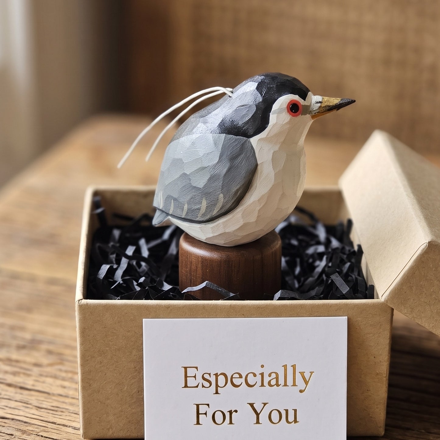 A carved Black-crowned Night Heron figurine with a geometric faceted pattern sits in an open kraft gift box filled with black paper shred. The box has a white card that reads 'Especially For You'.