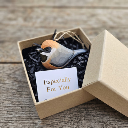 A small Black-throated bushtit bird-shaped ornament with a geometric faceted design in orange, black, white, and gray, presented in a kraft gift box with black shredded paper and a card that reads 'Especially For You' on a rustic wooden surface.