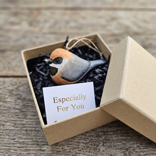 A small Black-throated bushtit bird-shaped ornament with a geometric faceted design in orange, black, white, and gray, presented in a kraft gift box with black shredded paper and a card that reads 'Especially For You' on a rustic wooden surface.