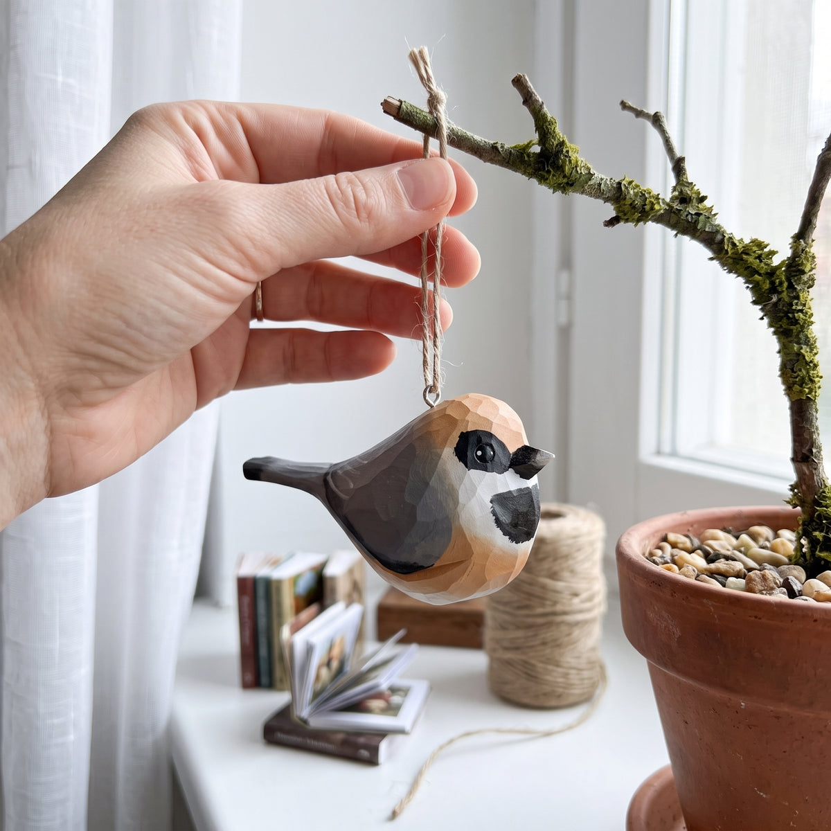 A hand holds a small painted Black-throated bushtit ornament in front of a window, with a potted plant and decorative books in the background.