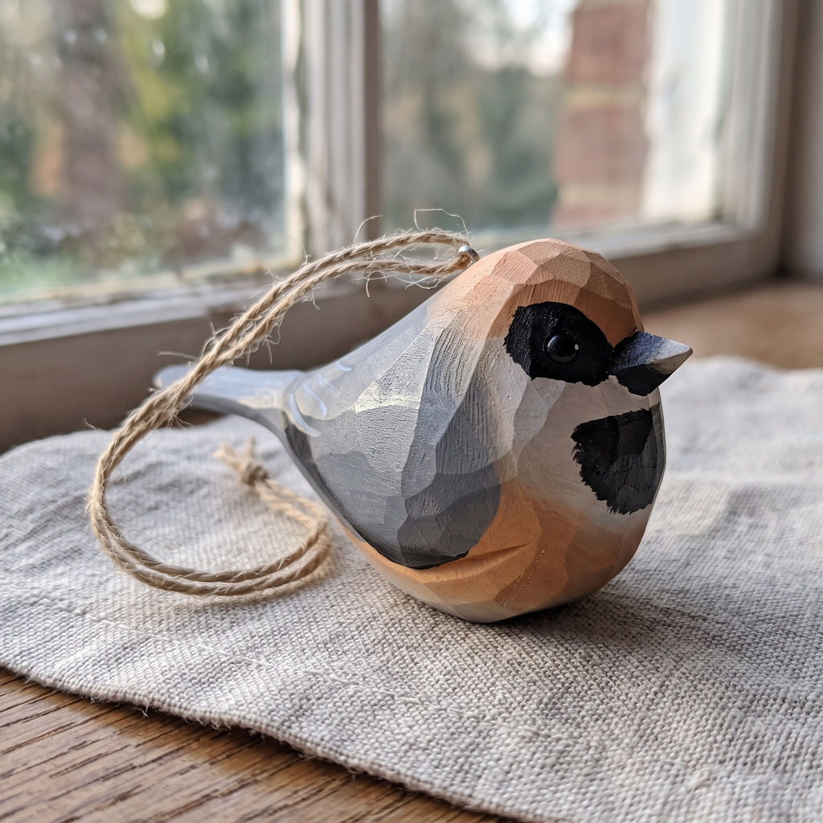 A handcrafted wooden Black-throated bushtit bird ornament with a natural twine loop, resting on a textured linen cloth near a window.