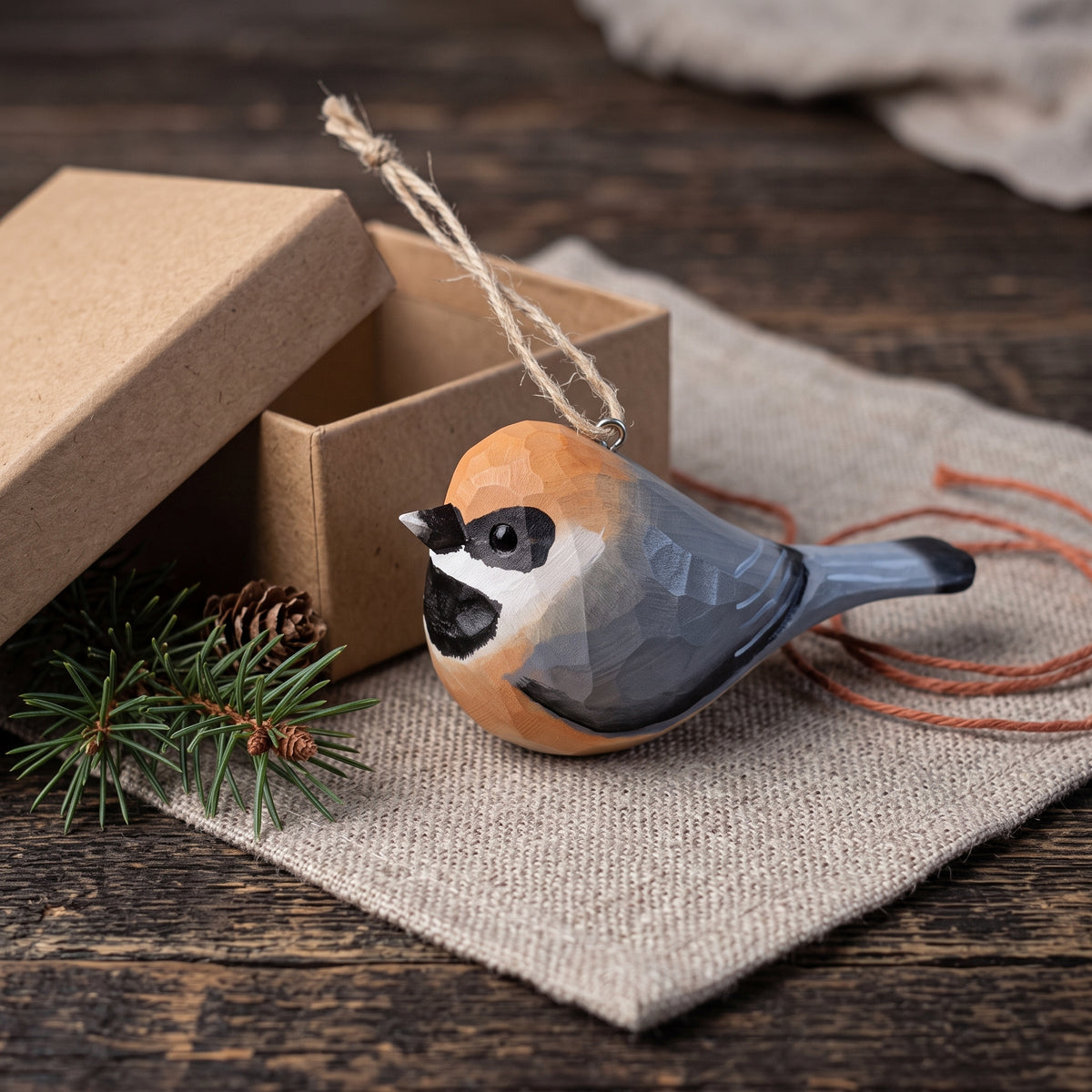 A carved wooden Black-throated bushtit bird ornament with a natural wood grain finish, featuring a black-capped chickadee design, resting on a burlap cloth next to its kraft paper gift box.