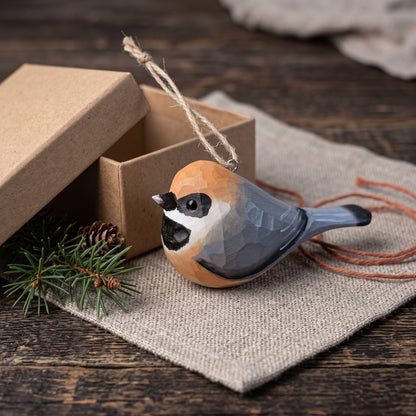 A carved wooden Black-throated bushtit bird ornament with a natural wood grain finish, featuring a black-capped chickadee design, resting on a burlap cloth next to its kraft paper gift box.