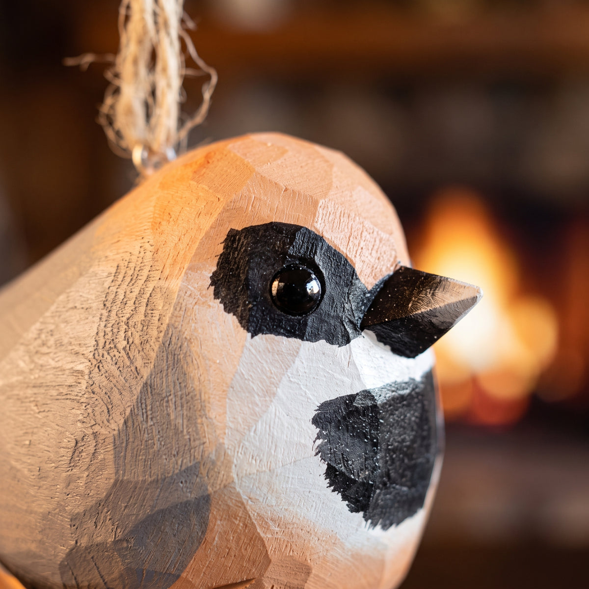A close-up of a hand-carved wooden Black-throated bushtit bird ornament, likely a chickadee, with a natural wood finish, black painted facial markings, and a hanging twine loop, set against a warm, blurred background of a fireplace.