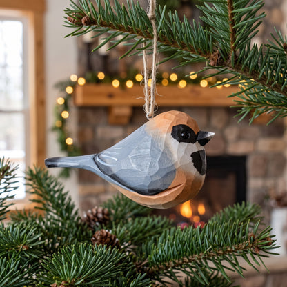 A geometric, low-poly style wooden Black-throated bushtit bird ornament, resembling a chickadee, hanging from a jute twine string on a pine tree branch in a cozy, blurred fireplace setting.