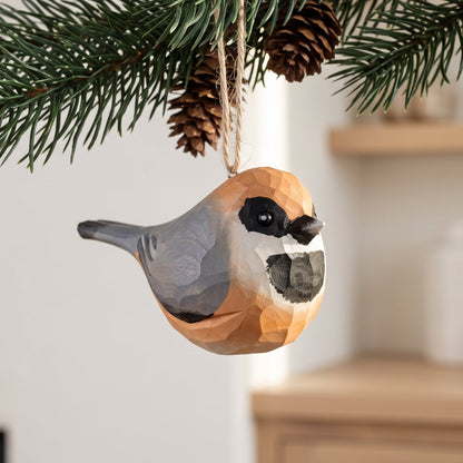 A geometric, low-poly Black-throated bushtit ornament hanging from a pine branch with pinecones in a cozy indoor setting.