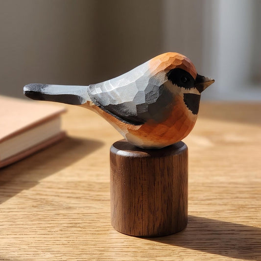 A hand-carved wooden Black-throated bushtit figurine with a geometric faceted finish, perched on a small cylindrical wooden base. The bird features a warm orange-brown back, a gray head and wings, and a black eye.