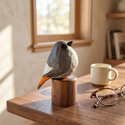 A geometric Black redstart sculpture made of faceted polygonal pieces sits on a wooden cylindrical base on a wooden table in a sunlit room. A cream ceramic mug and tortoiseshell eyeglasses are nearby, with a blurred window and shelf in the background.