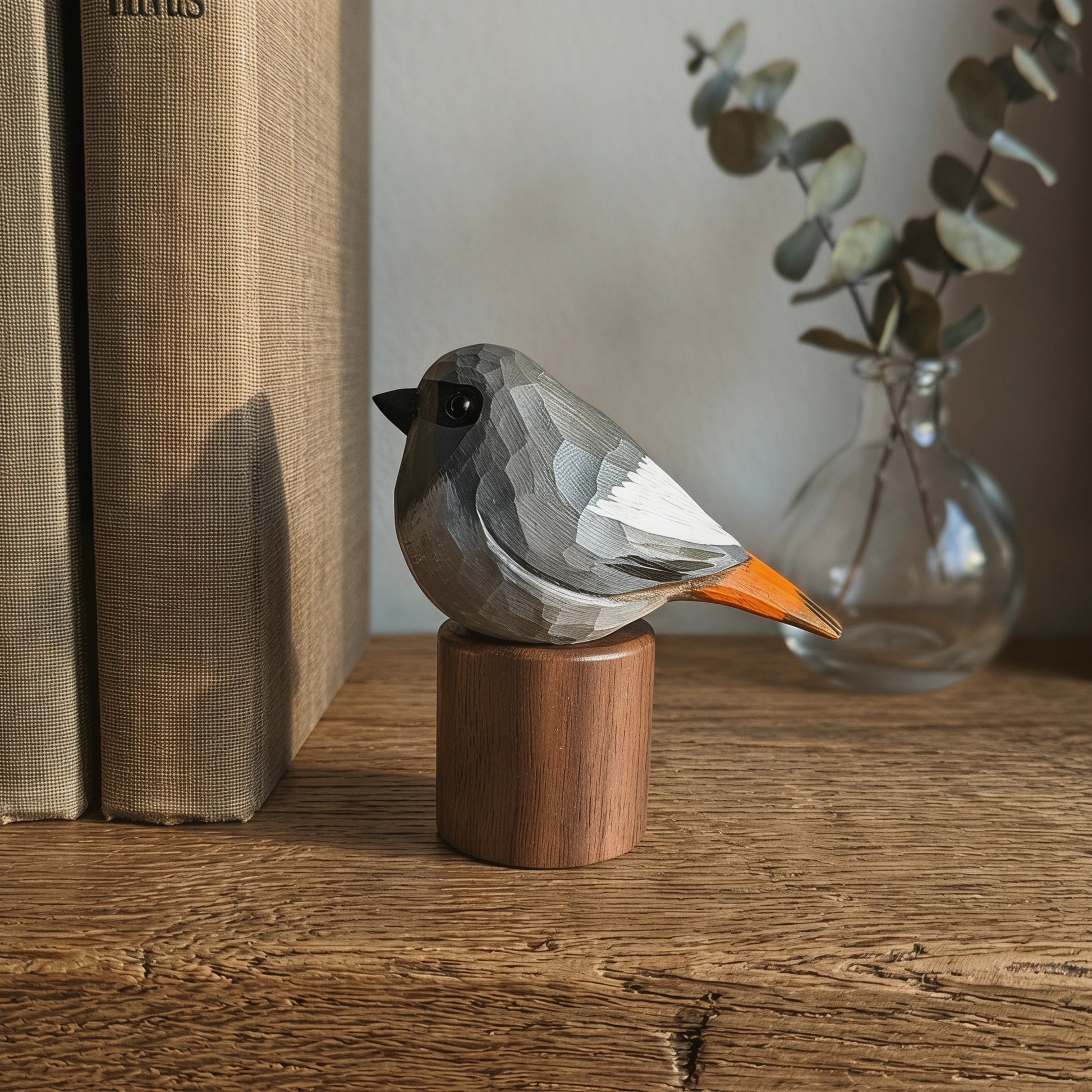 A geometric, faceted Black redstart figurine with a gray, white, and orange color scheme, perched on a small wooden cylinder. The figurine is styled on a rustic wooden table next to books and a glass vase with eucalyptus.