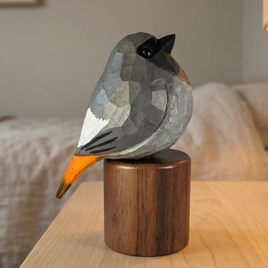 A geometric Black redstart figurine with a gray, white, and black faceted body and a bright orange beak, perched on a dark brown wooden cylinder. The figurine is displayed on a light wood bedside table next to a lit lamp in a softly blurred bedroom setting.