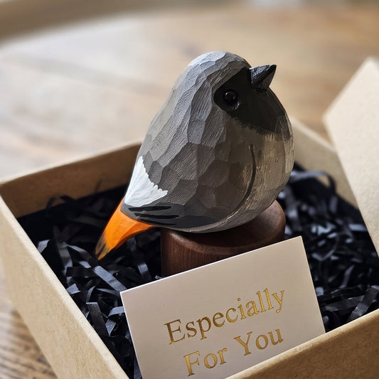 A small Black redstart figurine with a gray and black body, white wing, and orange beak, displayed inside a brown kraft gift box with black shredded paper and a card that reads 'Especially For You'.
