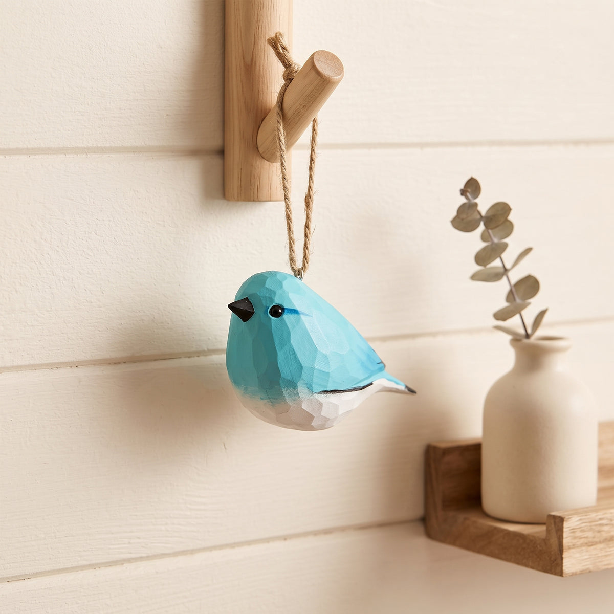 Hand-carved wooden bluebird ornament hanging from a wooden wall peg beside a ceramic vase with dried eucalyptus