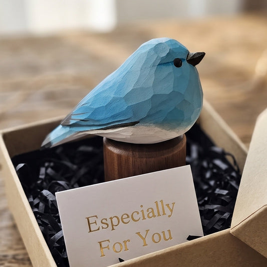 A small blue bird figurine displayed in an open kraft gift box with black shredded paper filler and a card that reads 'Especially For You.' The box sits on a rustic wooden surface with a blurred background.