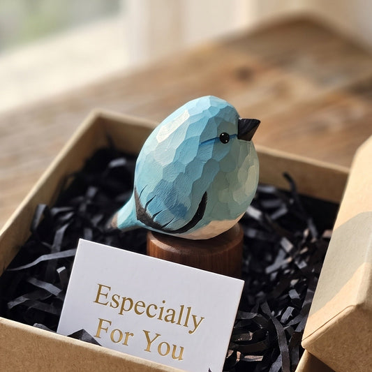 A small faceted blue bird figurine displayed in a kraft gift box with black shredded filler and a card that reads 'Especially For You,' set on a wooden tabletop near a window.