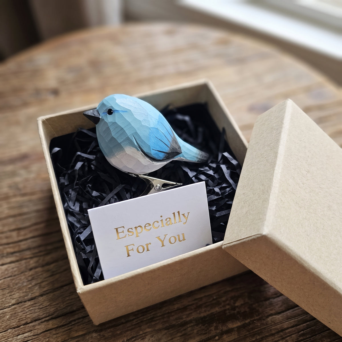 A hand-carved blue and white wooden bird ornament resting in an open kraft gift box filled with black shredded paper, alongside a small card reading "Especially For You".
