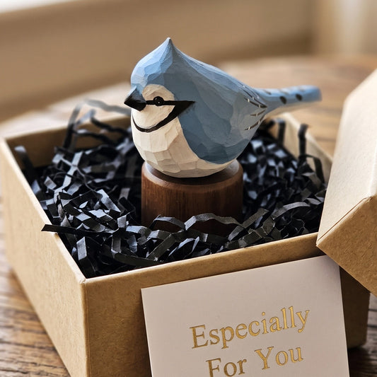 A small geometric Blue jay figurine in light blue and white sits in a brown kraft gift box filled with black paper shred, with a card reading 'Especially For You' in front. The box is open on a wooden surface in a softly lit indoor setting.