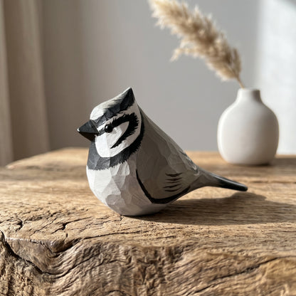 A low-poly wooden Bridled titmouse figurine in gray, black, and white, displayed on a rustic wooden table with a small white vase and dried grass in the background.