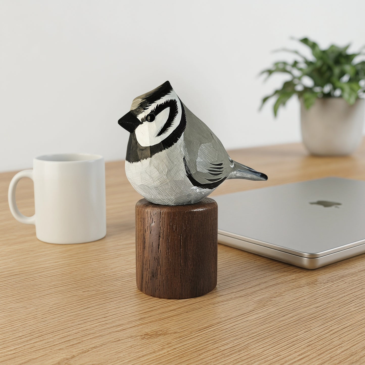 A hand-carved Bridled titmouse figurine perched on a wooden cylinder base, displayed on a light wood desk next to a white mug, a closed silver laptop, and a potted plant.