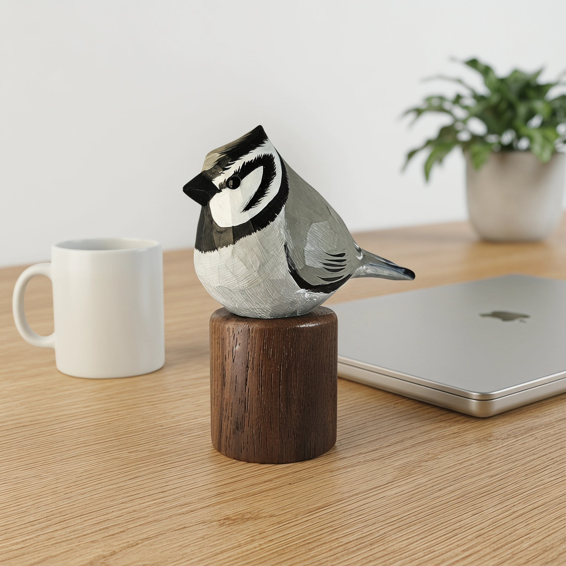 A hand-carved Bridled titmouse figurine perched on a wooden cylinder base, displayed on a light wood desk next to a white mug, a closed silver laptop, and a potted plant.