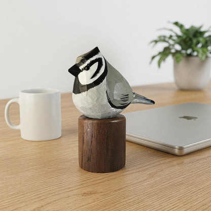 A hand-carved Bridled titmouse figurine perched on a wooden cylinder base, displayed on a light wood desk next to a white mug, a closed silver laptop, and a potted plant.