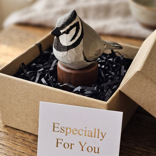 A small carved Bridled titmouse figurine displayed in an open kraft gift box with black shredded filler and a card reading 'Especially For You' in front. The bird has a gray, beige, and black carved wood finish and stands on a round wooden base.