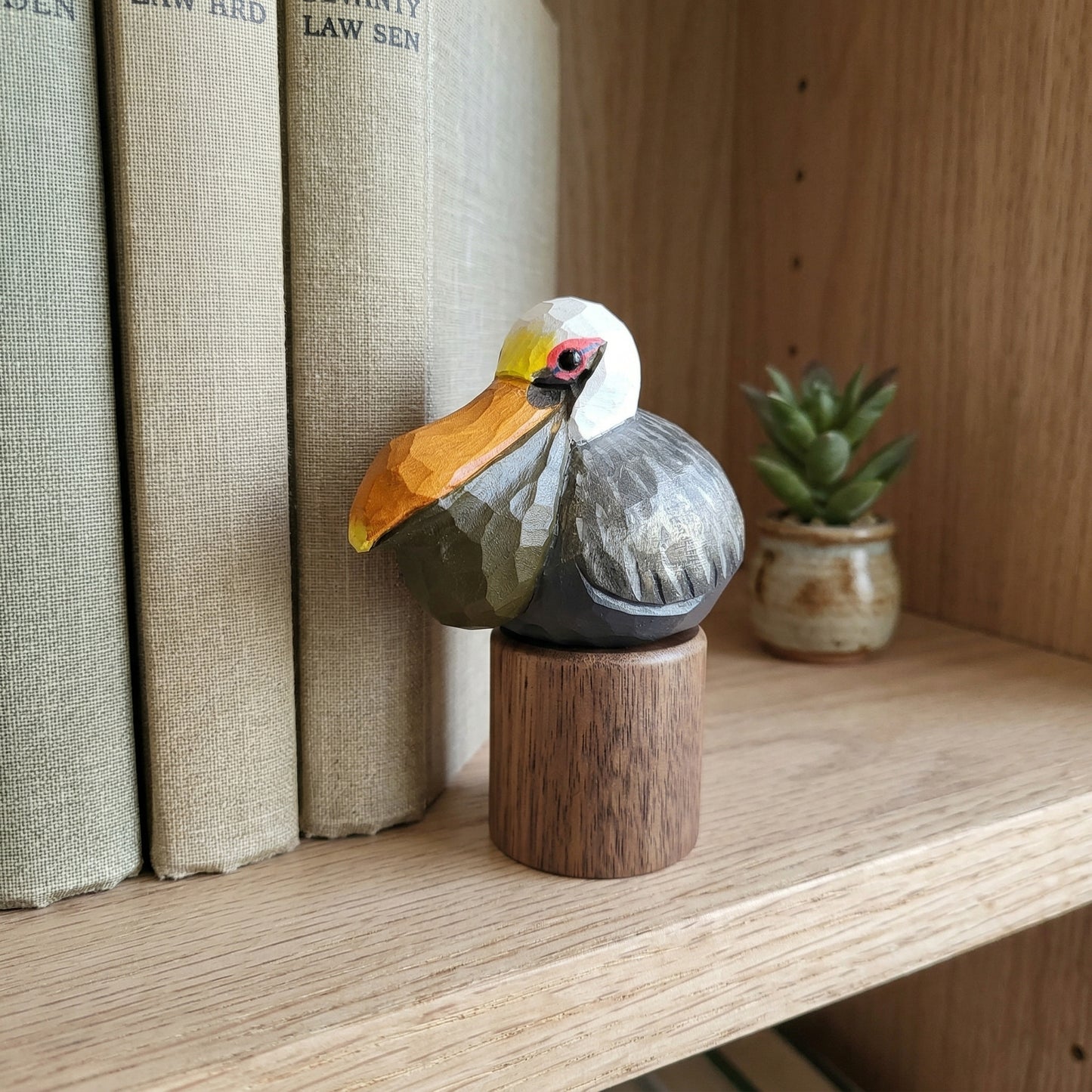 A carved wooden Brown pelican figurine with a white head, yellow beak, gray body, and red eye detail, mounted on a small round wooden base and displayed on a wooden shelf beside books and a small potted plant.