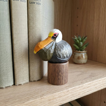 A carved wooden Brown pelican figurine with a white head, yellow beak, gray body, and red eye detail, mounted on a small round wooden base and displayed on a wooden shelf beside books and a small potted plant.