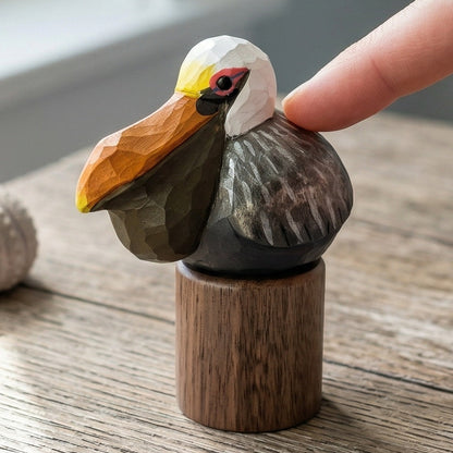 A hand is gently touching a small carved wooden Brown pelican figurine mounted on a cylindrical wooden base, placed on a rustic wooden table with a sea urchin and soft window light in the background.