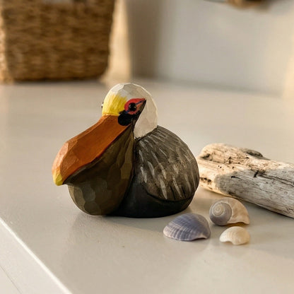 A hand-carved wooden Brown pelican figurine with a large orange beak, dark brown body, and white head detail, styled on a white tabletop with driftwood, seashells, and a woven basket in the background.