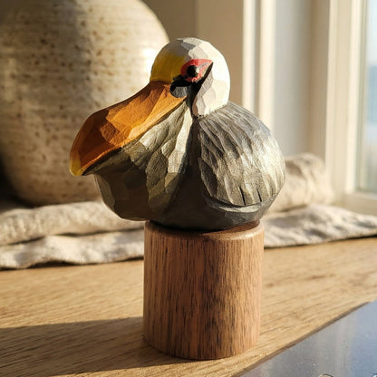 A hand-carved wooden Brown pelican figurine with a yellow beak and gray body, mounted on a small wooden base on a tabletop near a window.