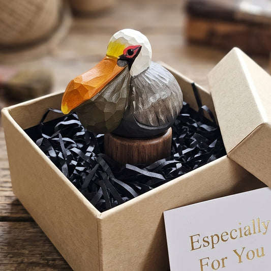 A small carved wooden Brown pelican figurine with a geometric, faceted orange beak and white head, presented in an open kraft gift box with black shredded paper filler and a card that reads 'Especially For You'. The background is a rustic wooden table with blurred craft supplies.