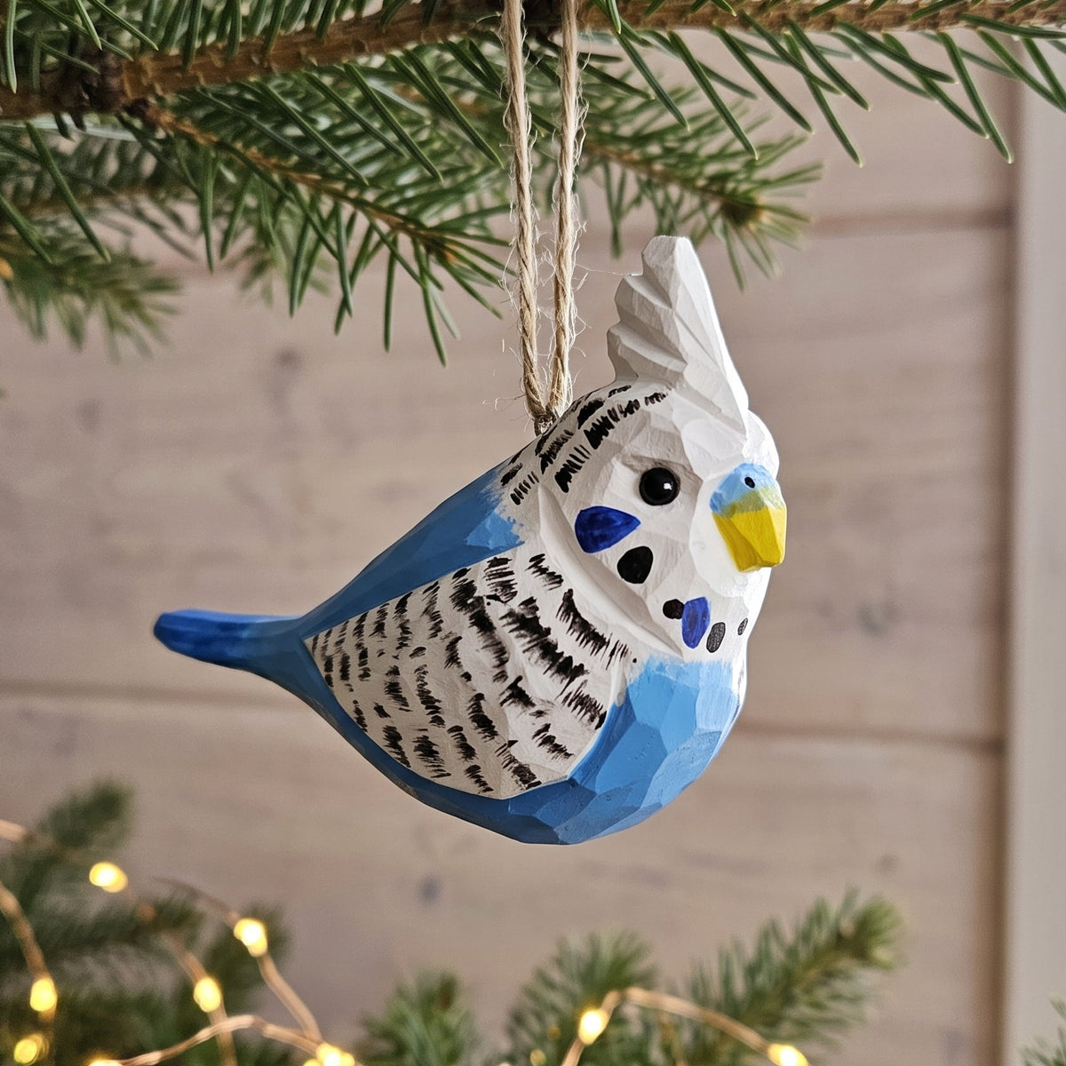 Hand-carved blue and white wooden budgie ornament hanging on a pine tree branch