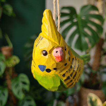 Close-up of yellow and green hand-carved wooden cockatiel budgie ornament hanging in front of an indoor plant