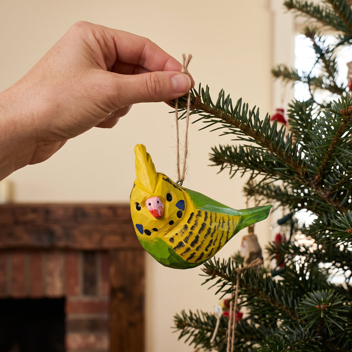 Hand hanging a yellow and green hand-carved wooden budgie ornament on a Christmas tree branch