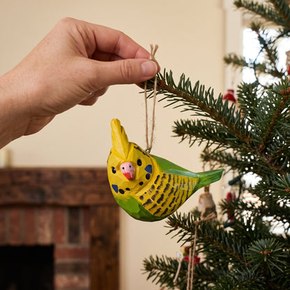 Hand hanging a yellow and green hand-carved wooden budgie ornament on a Christmas tree branch