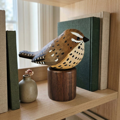 A hand-carved wooden Cactus wren figurine with brown, beige, and black spotted detailing, displayed on a dark wooden cylindrical base on a light wood shelf next to a small vase and books.
