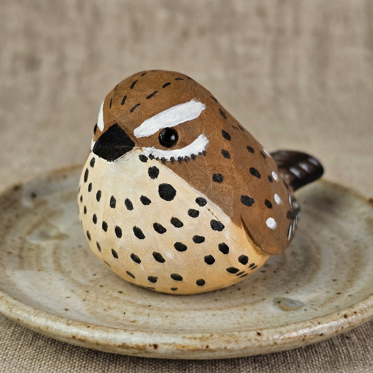 A small hand-painted wooden Cactus wren figurine with a speckled brown and beige body, resting on a rustic ceramic dish against a neutral burlap background.