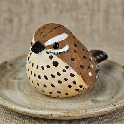 A small hand-painted wooden Cactus wren figurine with a speckled brown and beige body, resting on a rustic ceramic dish against a neutral burlap background.