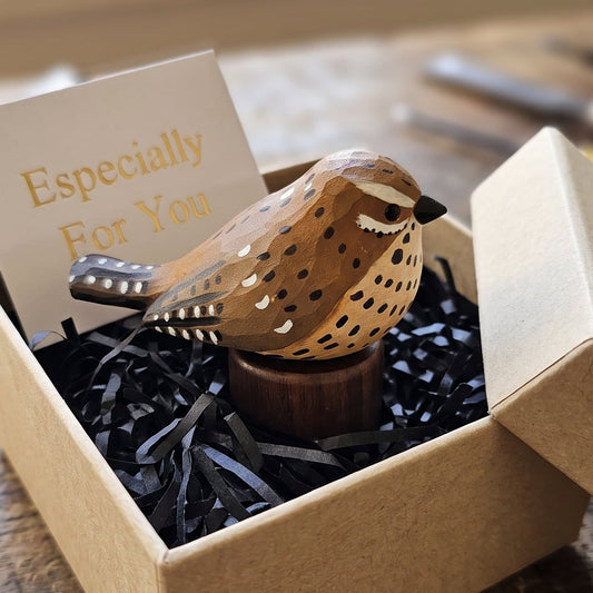 A small handcrafted wooden Cactus wren figurine with brown, beige, and black spotted detailing sits in a kraft gift box filled with black shredded paper and a card that reads 'Especially For You,' placed on a rustic wooden table.
